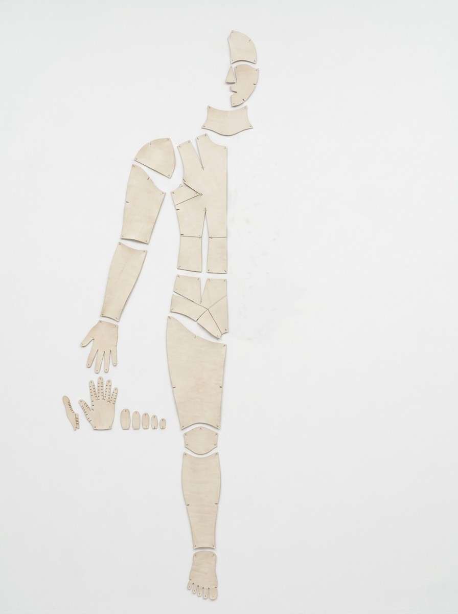 Pattern Pieces (Self): the left half of a life-size body pattern in flat cream kid leather, pinned to a white wall in anatomical position from head to foot, with the right half of the wall left empty.